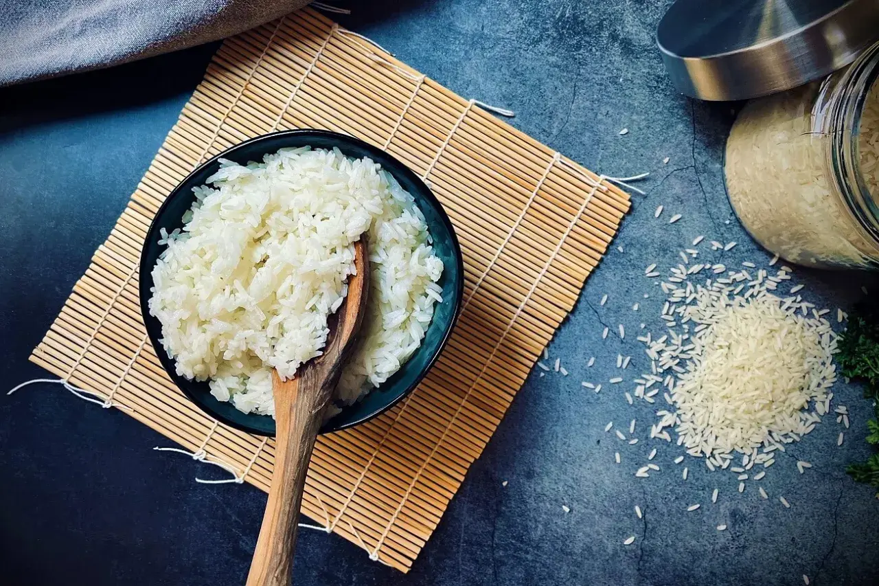 Schüssel mit gekochtem Reis auf Bambusmatte, daneben ungekochter Reis. Die Frage "wie lange muss Reis kochen" wird hier visuell beantwortet.