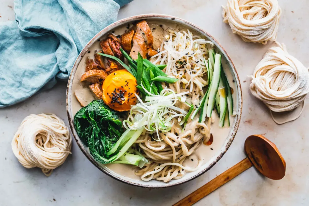 Schüssel mit cremiger Ramen-Suppe, Hühnchen, Ei, Gemüse und frischen **Rahmen Nudeln** daneben.