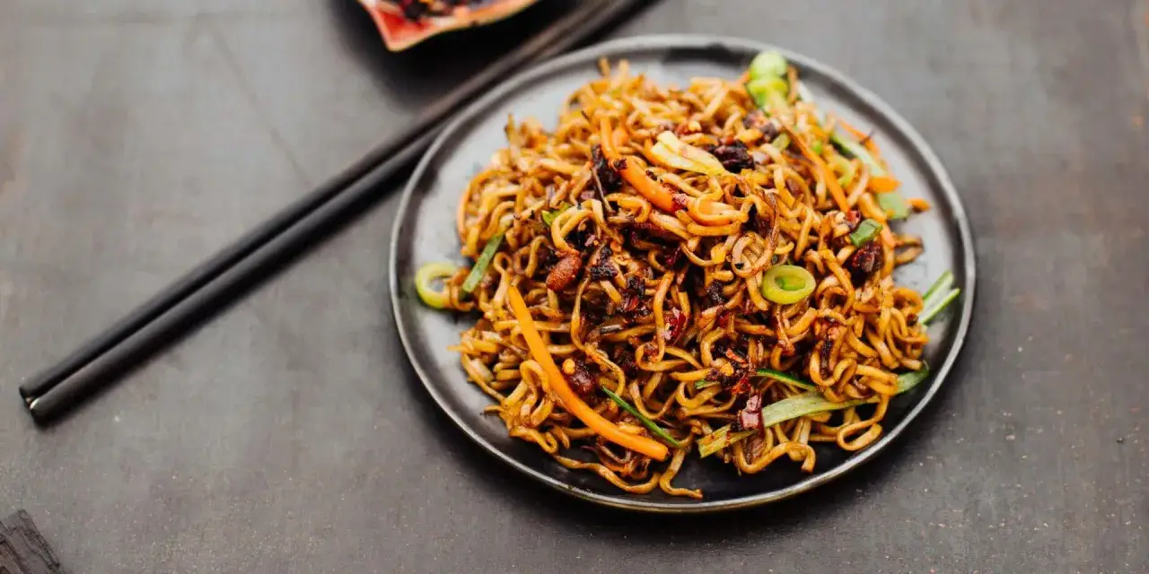 Ein Teller mit k&ouml;stlichem Chow Mein, garniert mit Fr&uuml;hlingszwiebeln und Karotten. Daneben liegen Essst&auml;bchen bereit.