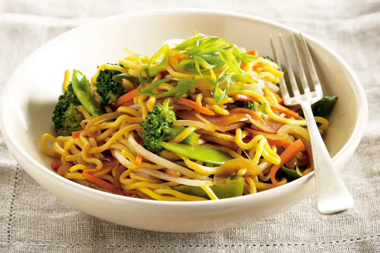 Sch&uuml;ssel mit chinesischen Eiernudeln, garniert mit Fr&uuml;hlingszwiebeln, Brokkoli, Karotten und Zuckerschoten. Eine Gabel liegt daneben.