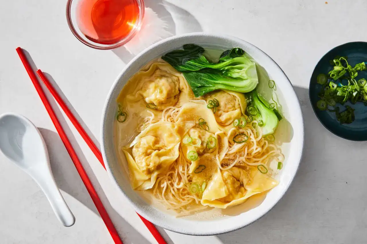 Eine Schüssel mit Wantan Suppe, Nudeln und Pak Choi. Daneben rote Stäbchen und eine Löffel.