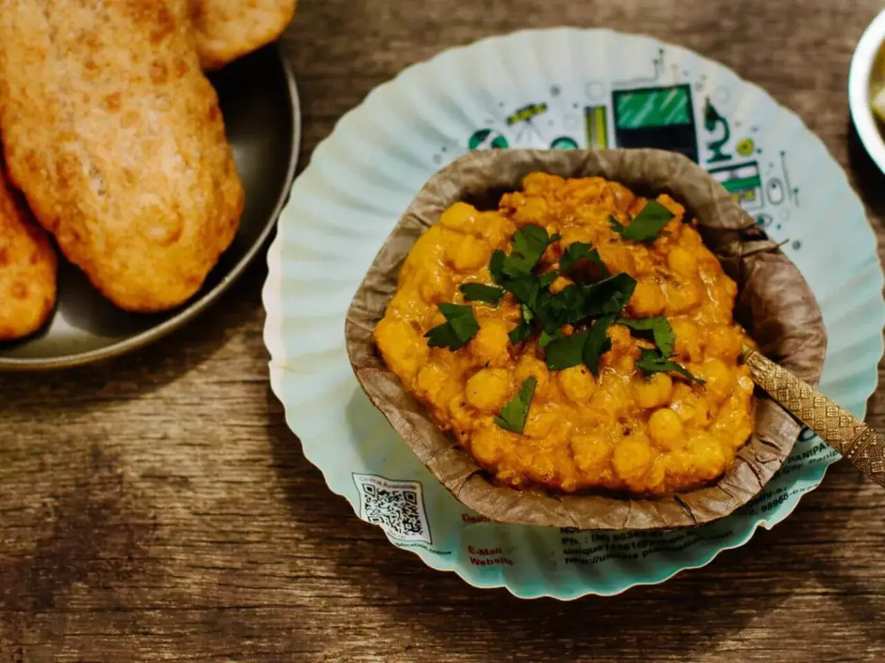 Ein Teller mit Chana Masala, garniert mit Koriander, neben knusprigem Bhatura Brot auf einem Holztisch.
