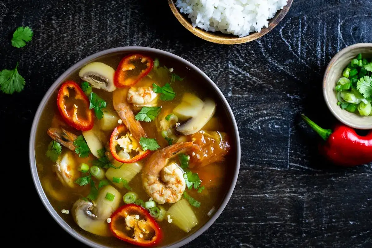 Eine Sch&uuml;ssel mit Canh Chua, einer vietnamesischen Suppe mit Garnelen, Pilzen und Chili, daneben Reis und Fr&uuml;hlingszwiebeln.