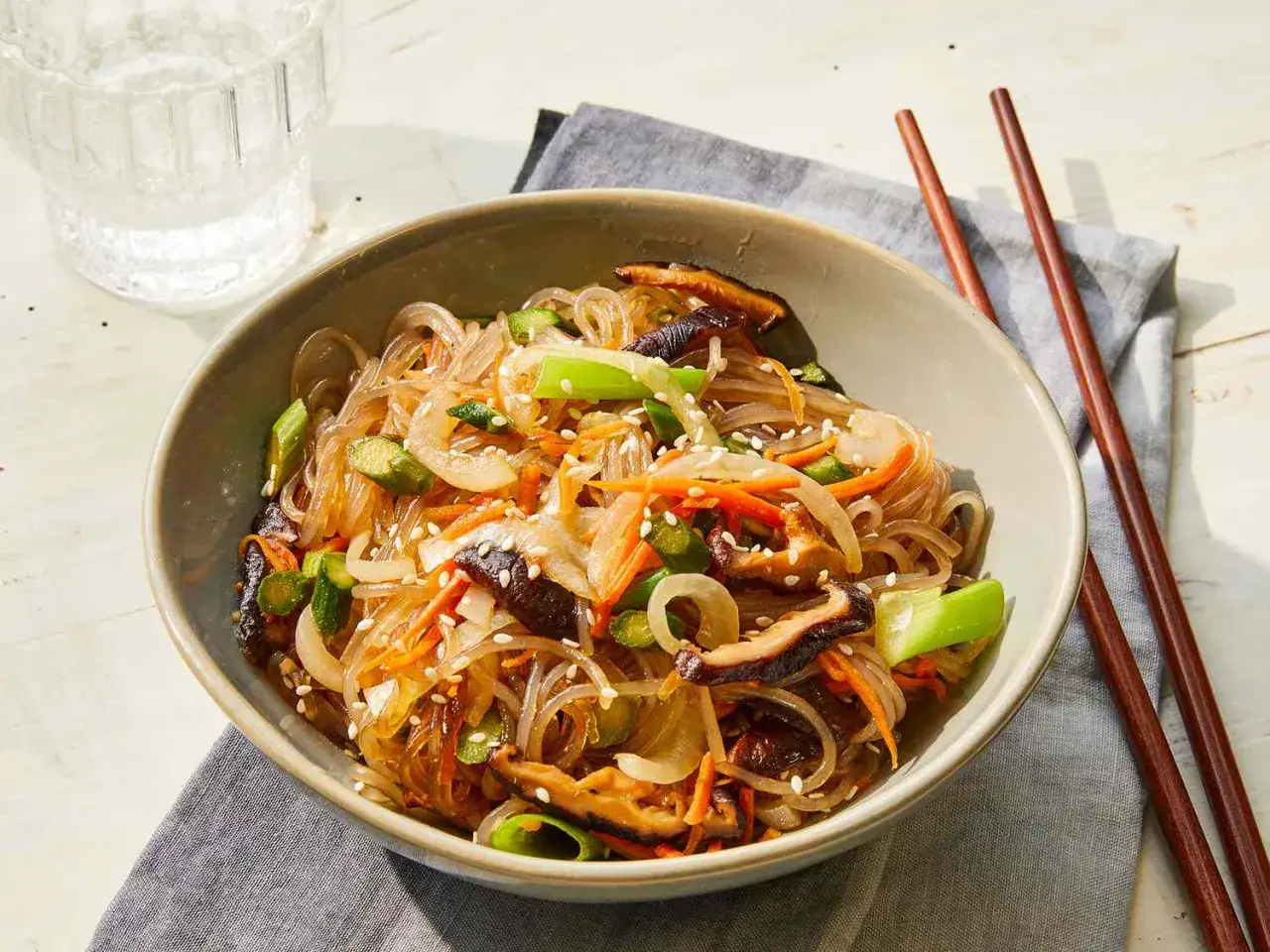 Sch&uuml;ssel mit Japchae, einem koreanischen Nudelgericht mit Glasnudeln, Gem&uuml;se und Pilzen, garniert mit Sesam. Ein Glas Wasser und St&auml;bchen daneben.
