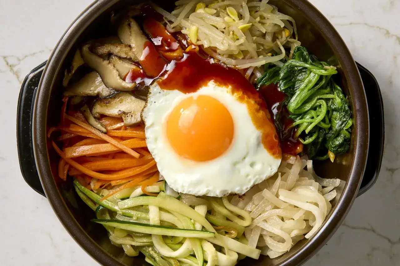 Sch&uuml;ssel mit Bibimbap: Reis, gebratenes Ei, Pilze, Karotten, Zucchini, Spinat und Sojasprossen, garniert mit scharfer So&szlig;e. Ein k&ouml;stliches Bibimbap Rezept!