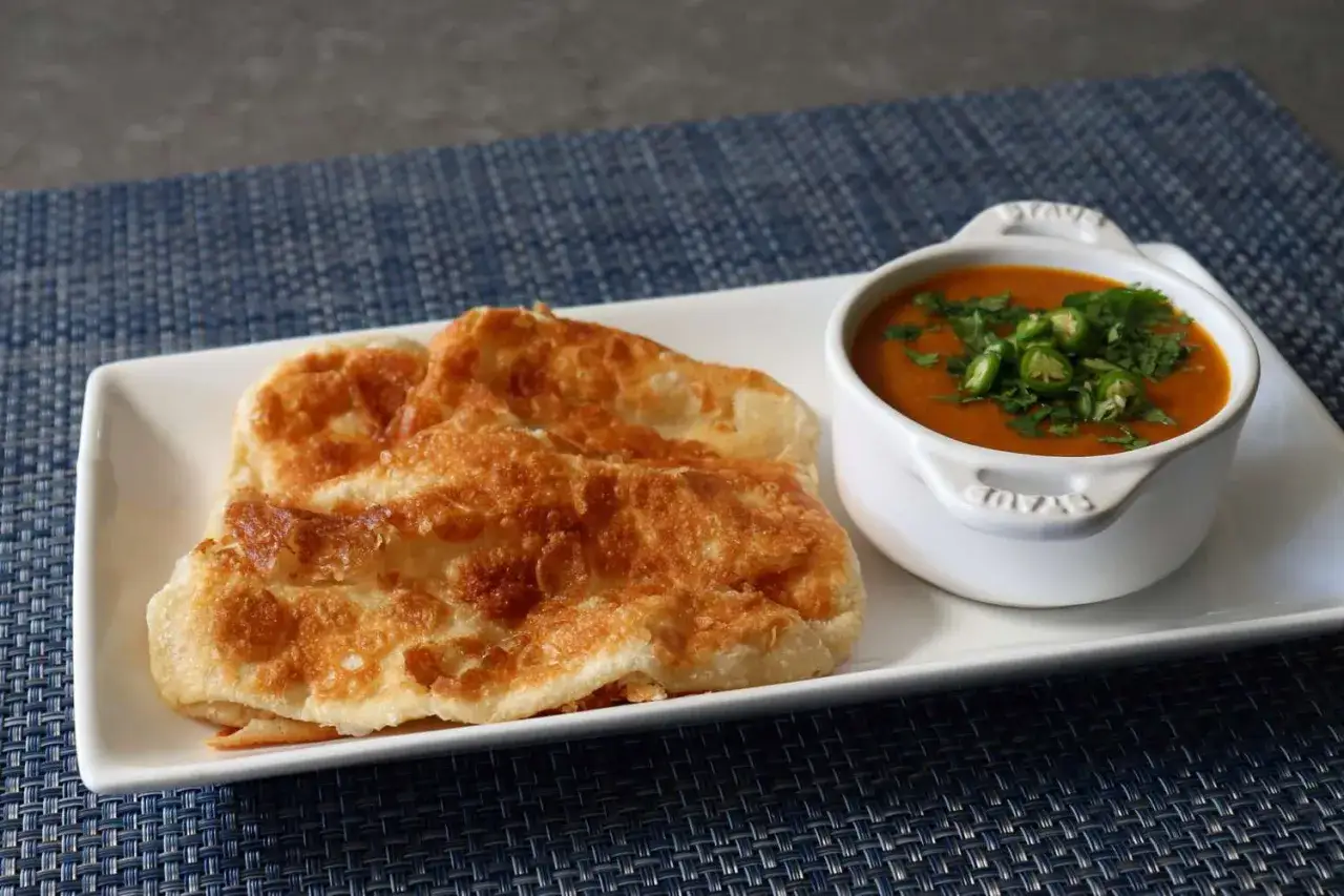 Goldbraunes, knuspriges Roti Canai liegt neben einer Sch&uuml;ssel mit w&uuml;rzigem Curry, garniert mit gr&uuml;nen Chilis und Koriander.