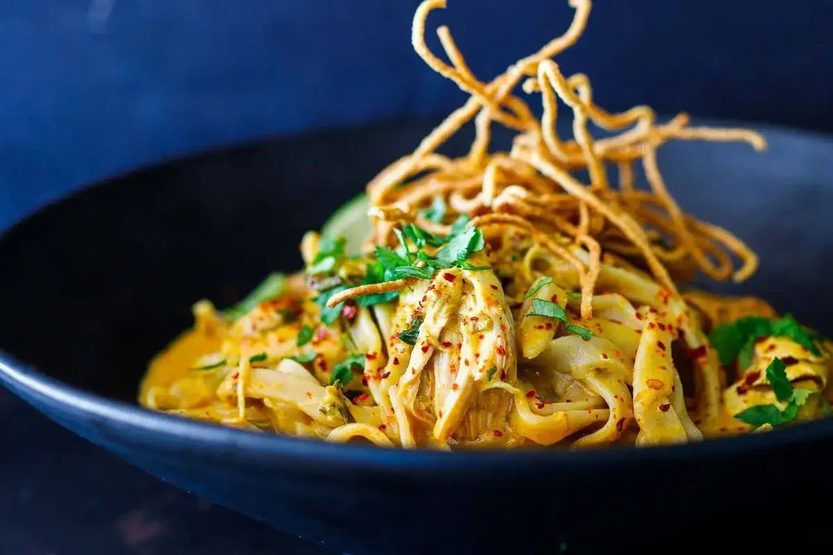 Khao Soi: cremige Kokosmilch-Currysauce mit zarten H&uuml;hnchenstreifen, weichen Nudeln und knusprigen Toppings in einer schwarzen Sch&uuml;ssel.