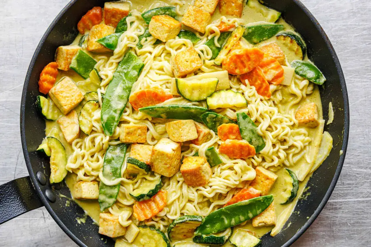 Eine Pfanne voller cremiger lo mein nudeln mit gebratenem Tofu, Zucchini, Karotten und Zuckerschoten in einer gr&uuml;nen Curry-Sauce.
