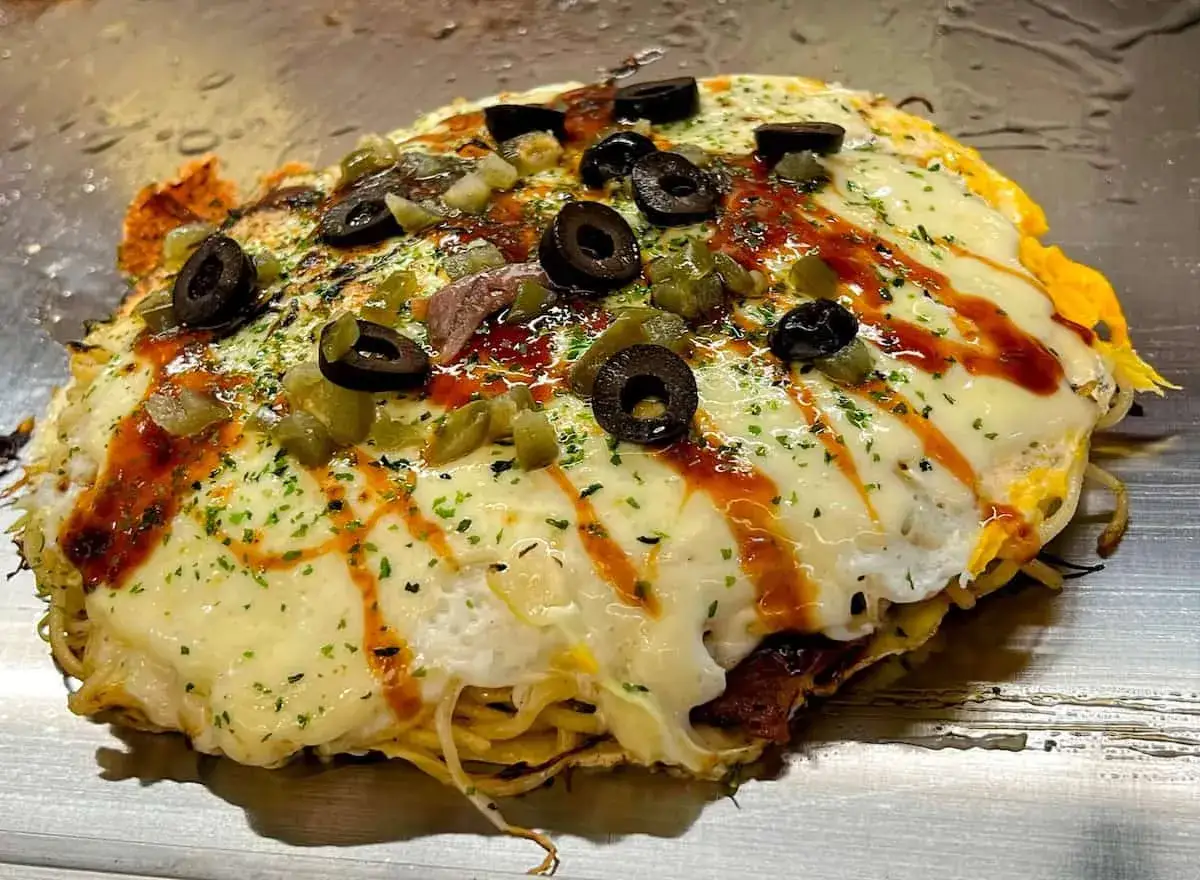 Ein Okonomiyaki im Hiroshima-Stil, überbacken mit geschmolzenem Käse, Oliven und würzigen Toppings.