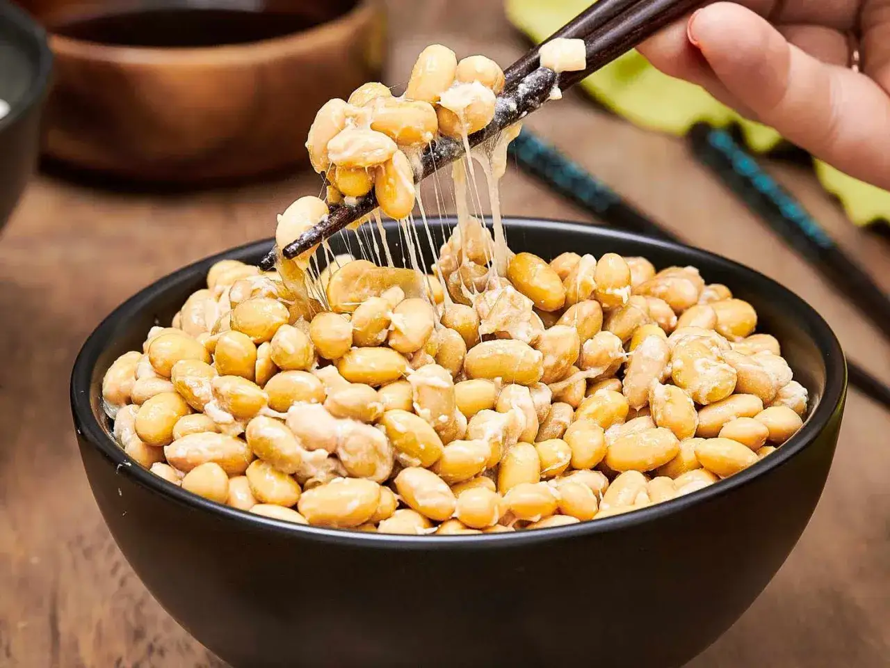 Fermentierte Sojabohnen, bekannt als Natto, werden mit St&auml;bchen aus einer schwarzen Sch&uuml;ssel gehoben. Die klebrige Textur ist deutlich sichtbar.