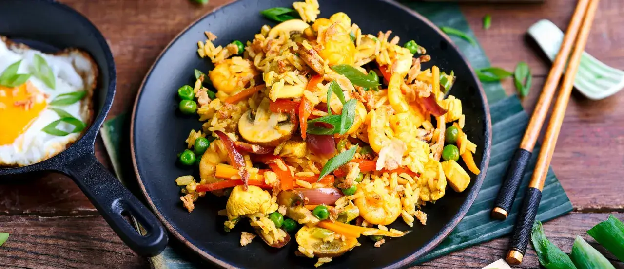 Nasi Goreng mit Garnelen, Pilzen und Erbsen auf einem schwarzen Teller, daneben ein Spiegelei in einer Pfanne und St&auml;bchen.