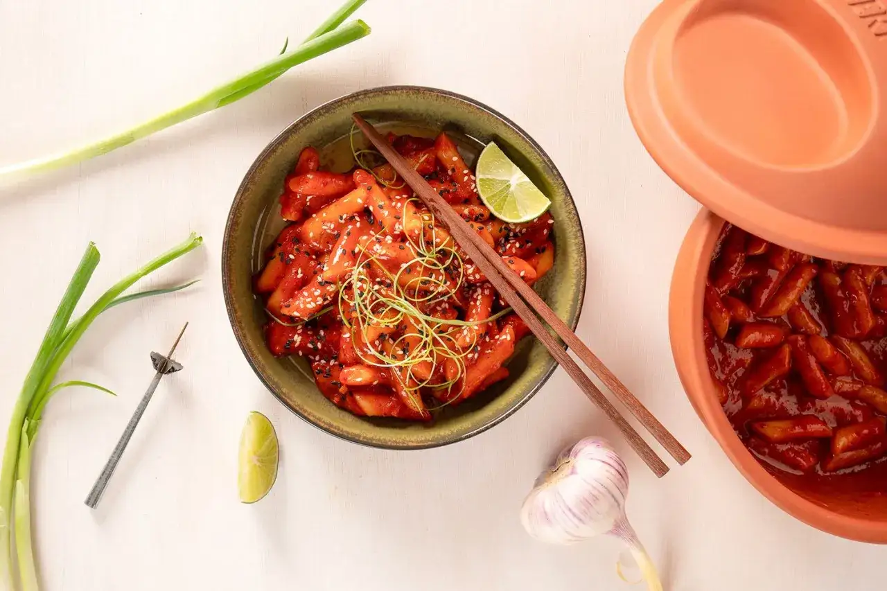 Schüssel mit würzigem Reiskuchen Tteokbokki, garniert mit Frühlingszwiebeln und Sesam. Daneben ein Tontopf mit mehr Tteokbokki.