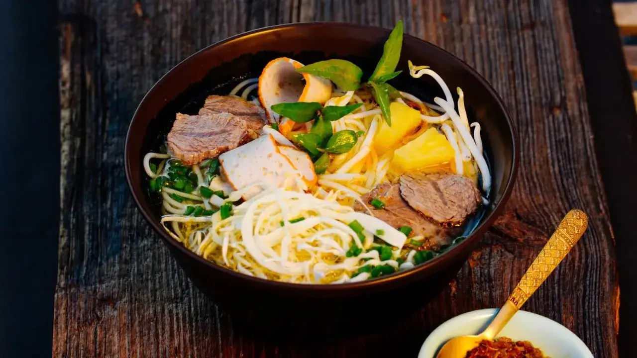 Herzhaftes Bun Bo Hue mit Rindfleisch, Ananasstücken, Kräutern und Sprossen in einer dunklen Schüssel auf Holzuntergrund.
