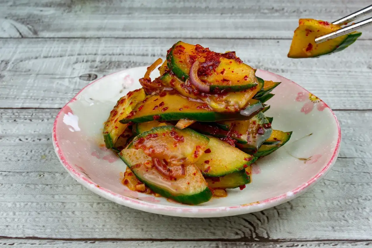 Ein Teller mit w&uuml;rzigem koreanischem Gurkensalat, der mit Chili und Zwiebeln mariniert ist. Ein St&uuml;ck wird mit St&auml;bchen angehoben.