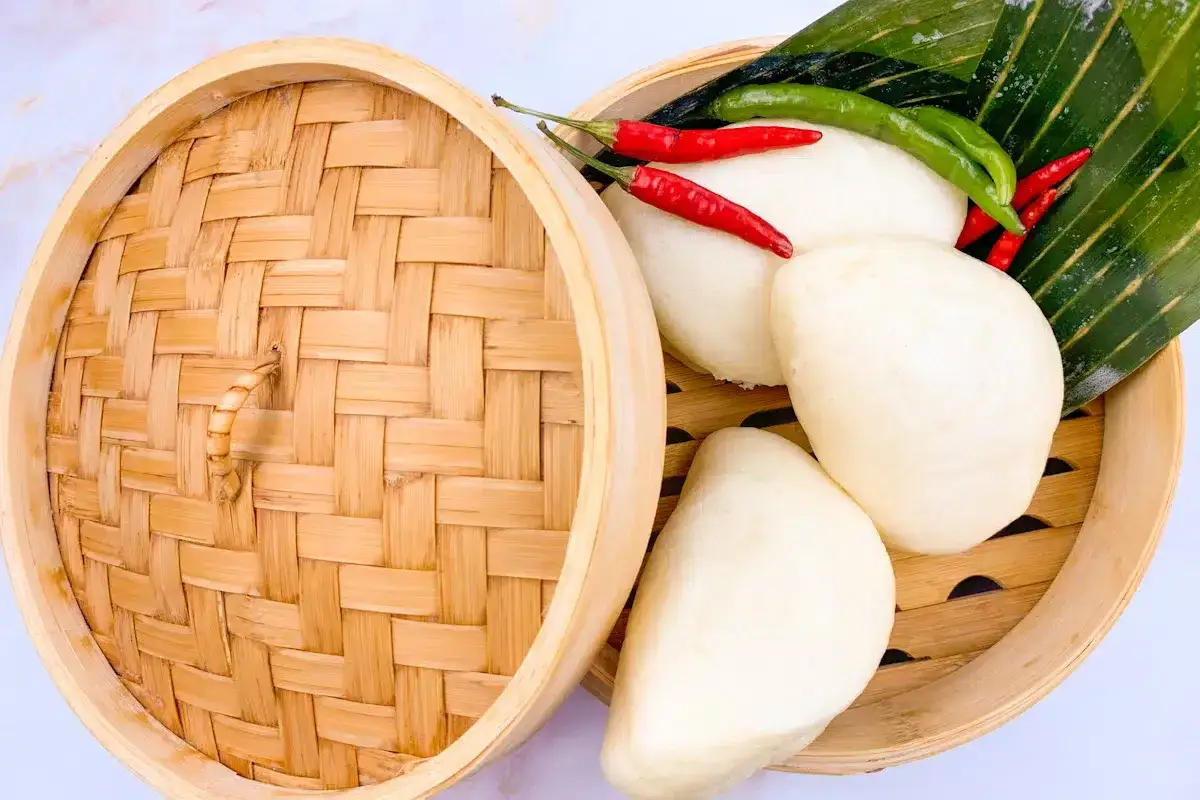 Dampfendes chinesisches Brot in einem Bambuskorb, garniert mit roten und gr&uuml;nen Chilis und einem gr&uuml;nen Blatt.