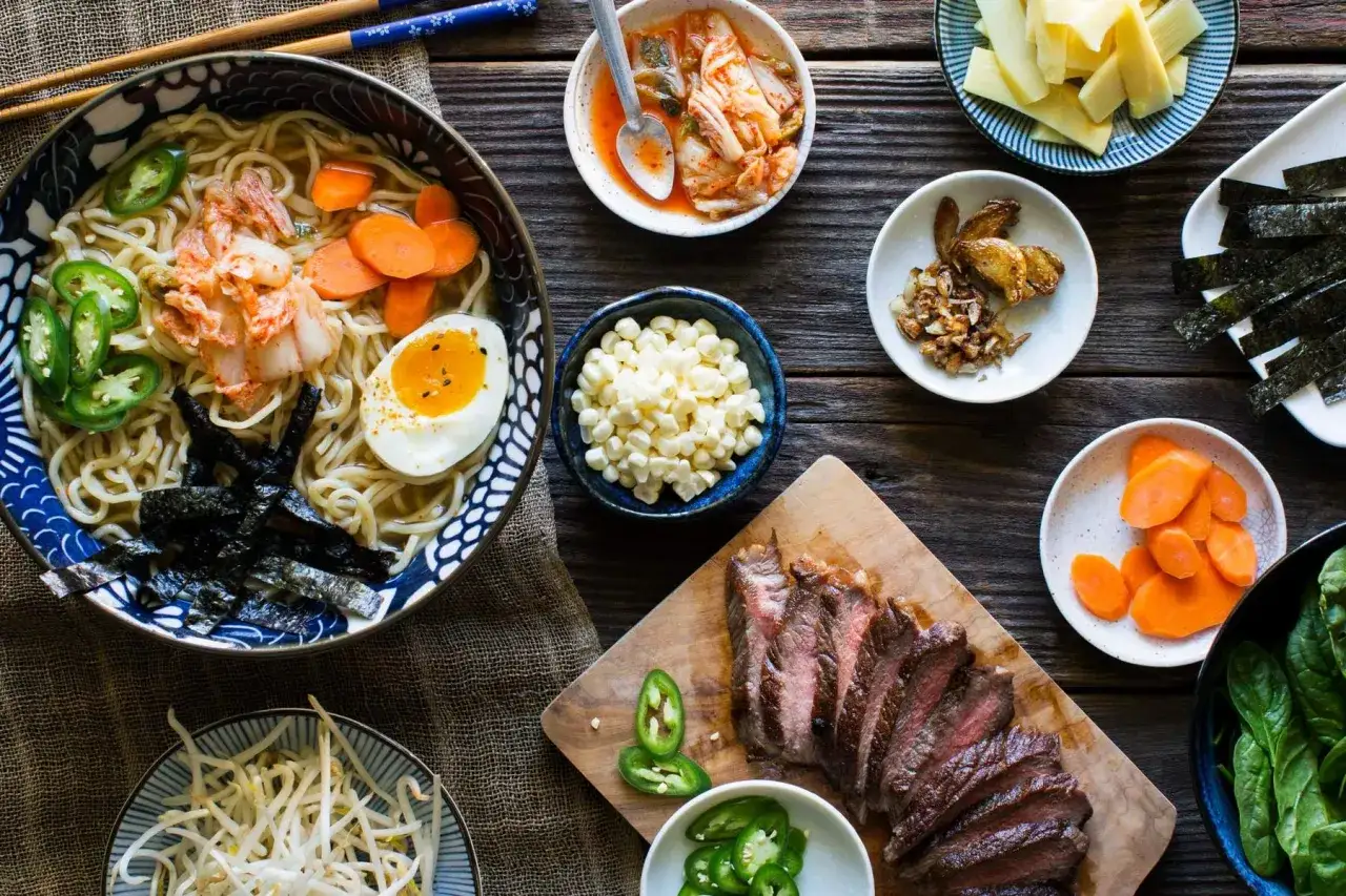 Sch&uuml;ssel mit Ramen, garniert mit Kimchi, Ei, Jalape&ntilde;os, Karotten und Nori. Daneben: Rindfleisch, Mais, Sprossen und weitere Ramen-Toppings.