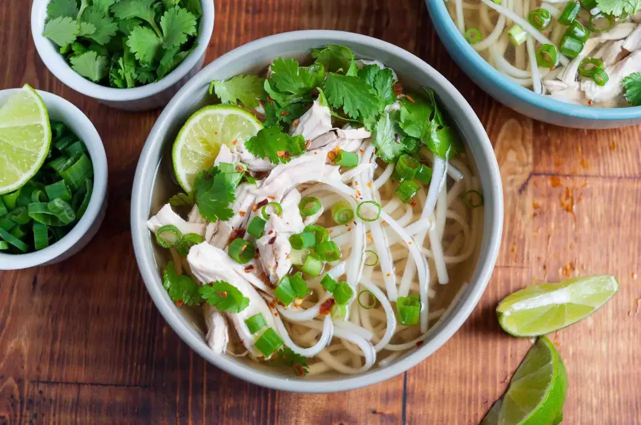Eine Sch&uuml;ssel dampfendes Pho Ga mit zarten H&uuml;hnerstreifen, Reisnudeln, Koriander, Fr&uuml;hlingszwiebeln und Limettenspalten.