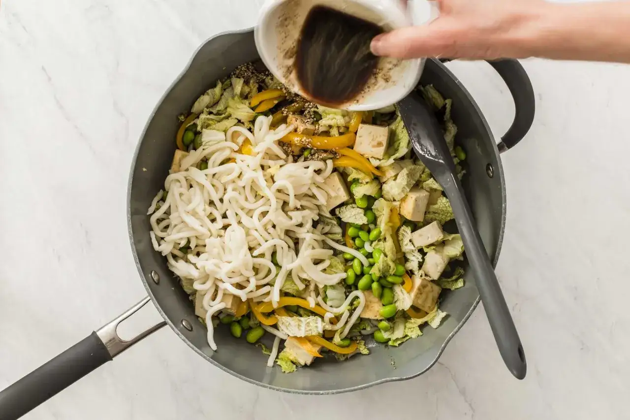 Wok mit Nudeln, Tofu, Gem&uuml;se und Edamame Proteinen. Eine Hand gie&szlig;t So&szlig;e dar&uuml;ber.