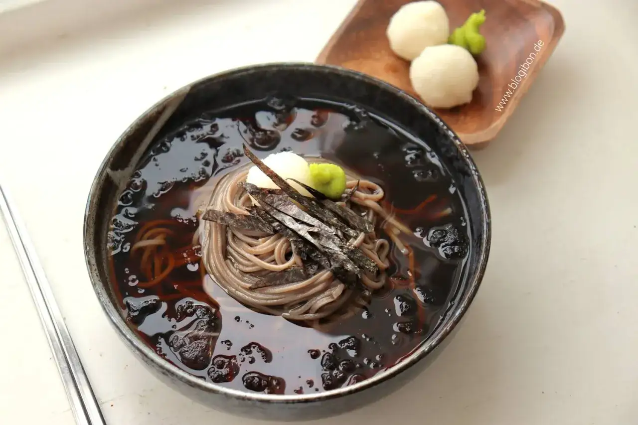 Eine Sch&uuml;ssel mit Mul Naengmyeon, kalter Buchweizennudelsuppe, garniert mit Algen und Wasabi. Daneben kleine Reisb&auml;llchen.