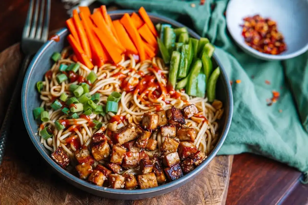 Sch&uuml;ssel mit asiatischen Nudeln, gebratenem Tofu, Karotten- und Paprikastreifen, garniert mit Fr&uuml;hlingszwiebeln und Chilisauce. Perfekt zum asiatische nudeln kochen.