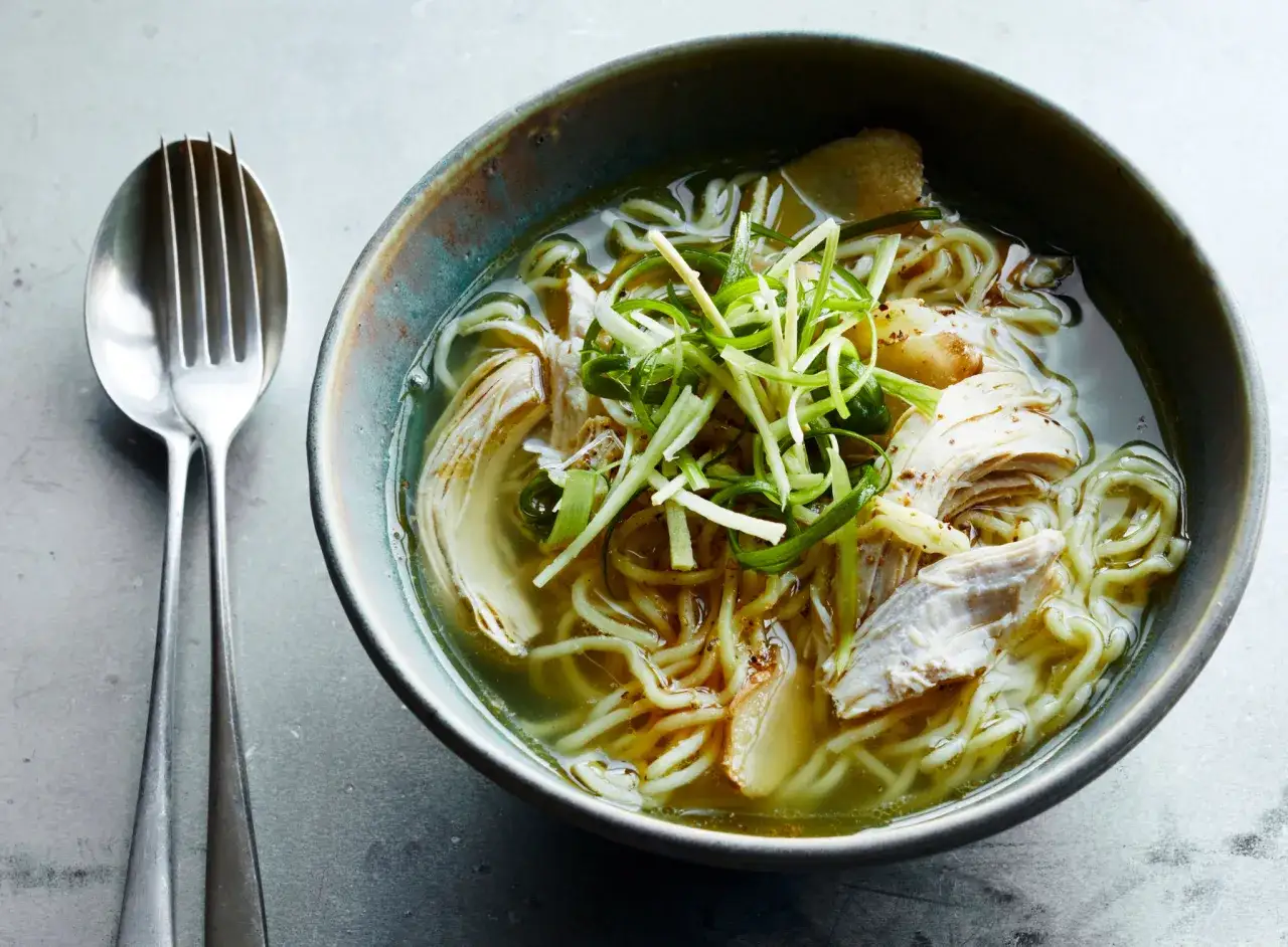 Eine dampfende Schüssel chinesische Suppe mit Huhn, Nudeln und Frühlingszwiebeln. Daneben liegen ein Löffel und eine Gabel.