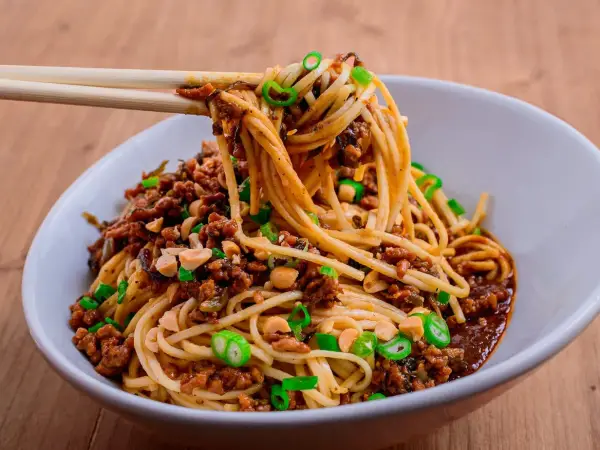 Ein Teller Dan Dan Noodles mit Hackfleisch, Erdnüssen und Frühlingszwiebeln, serviert mit Stäbchen.